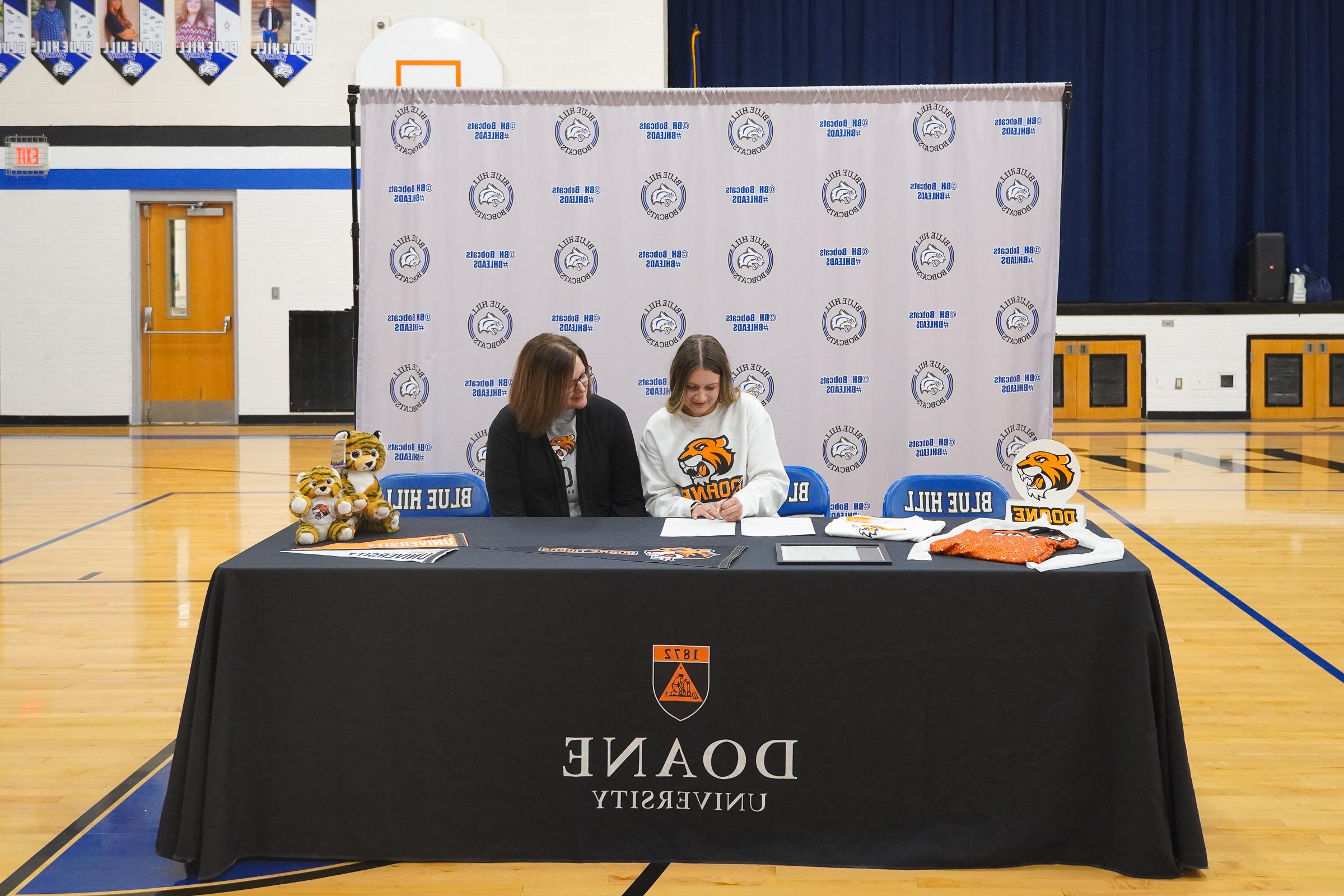 Two women sit behind a table draped with a black cloth that reads "澳门威尼斯人网址." The woman on the left wears a Doane sweatshirt and is signing a document. 他们周围是Doane的swag, 包括两只毛绒老虎, 几个三角旗, a shirt and a uniform for the Doane dance team.