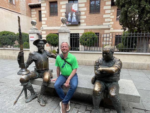 Josh Pope, wearing a green polo and jeans, sits between bronze statues of Sancho Panza and Don Quixote while traveling abroad in Madrid, Spain. Behind him is a brick building surrounded by a wrought-iron fence.