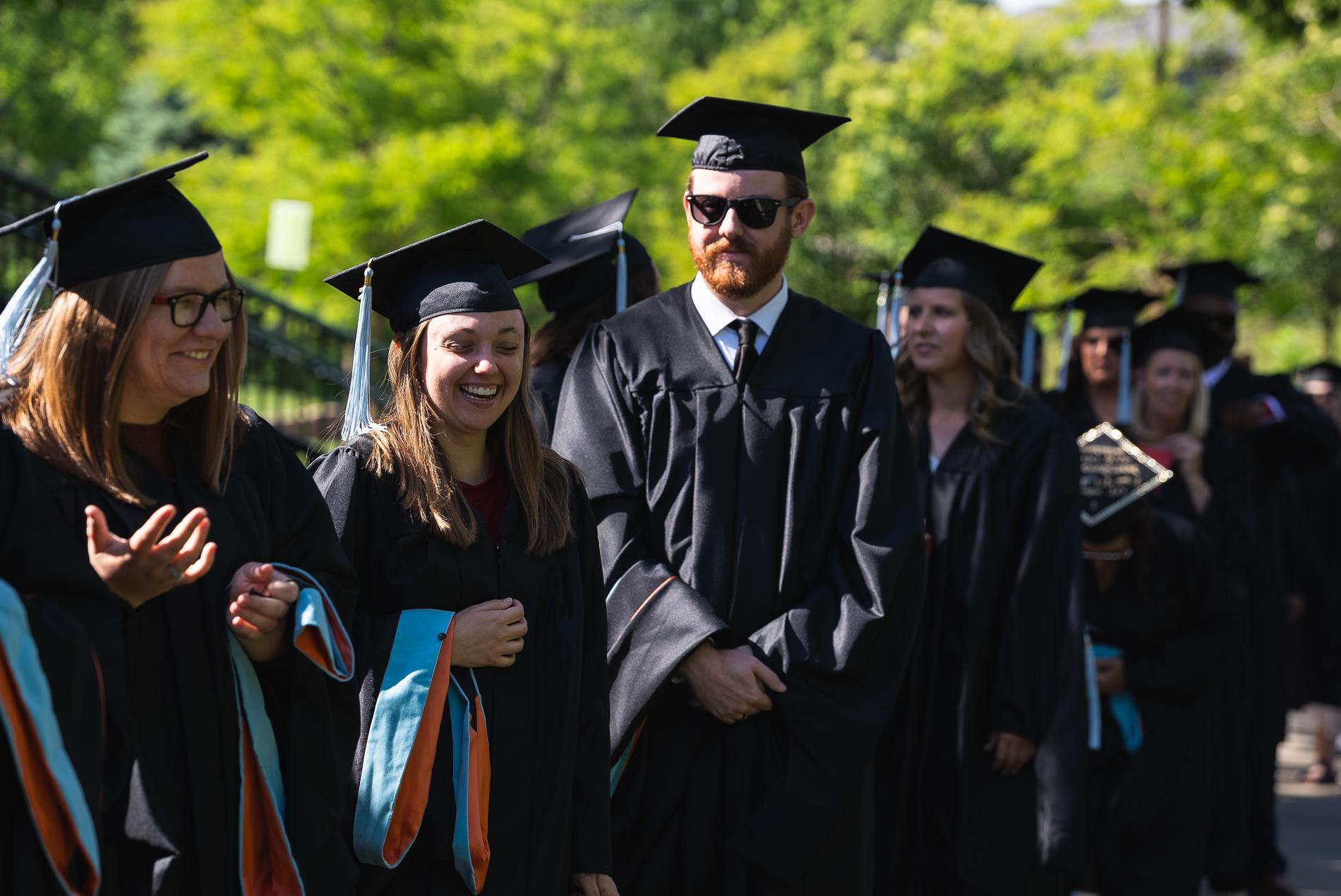 Graduates in black gowns 和 caps, with blue tassels, laugh 和 smile while lining up for commencement.