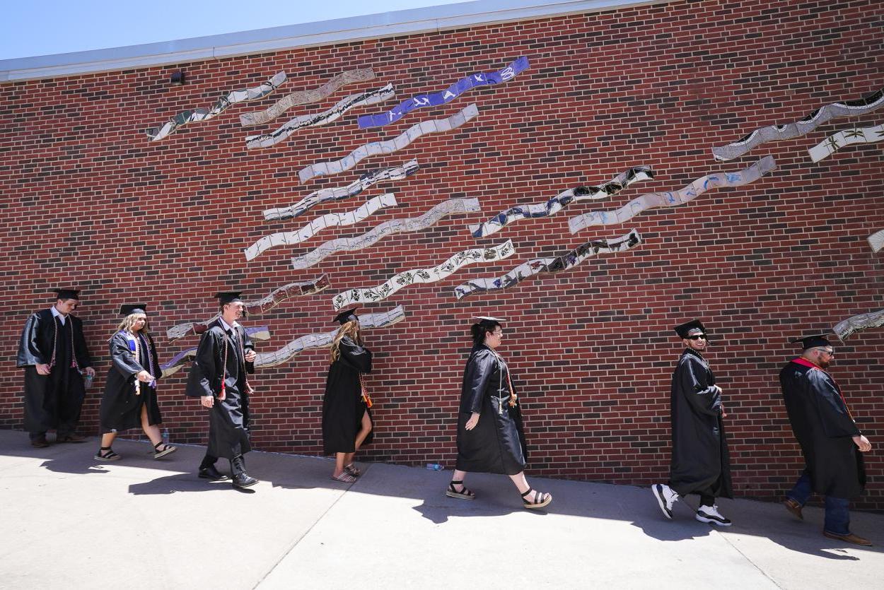 Seven students in black caps 和 gowns walk down a ramp in front of a brick wall showing a mural made of wavy tile.