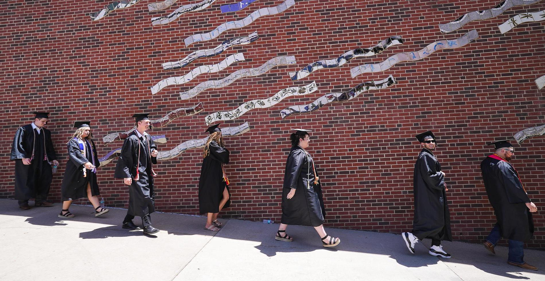 Seven students in black caps 和 gowns walk down a ramp in front of a brick wall showing a mural made of wavy tile.